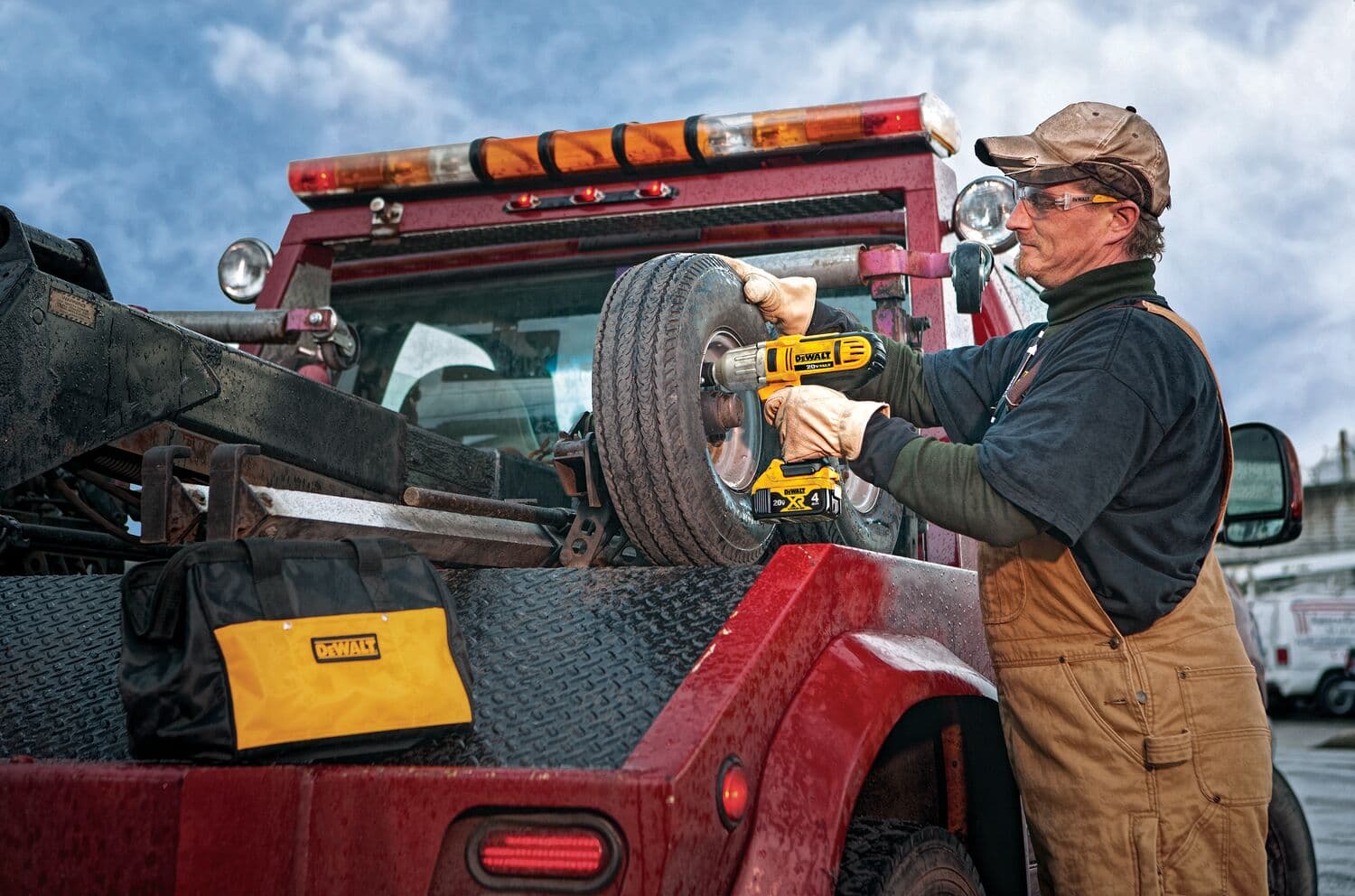 Una persona utiliza una llave de impacto inalámbrica DEWALT para aflojar o apretar los pernos de la rueda de un vehículo montada en una grúa roja. Una bolsa de herramientas amarilla y negra DEWALT está colocada en la plataforma del camión junto a la rueda.