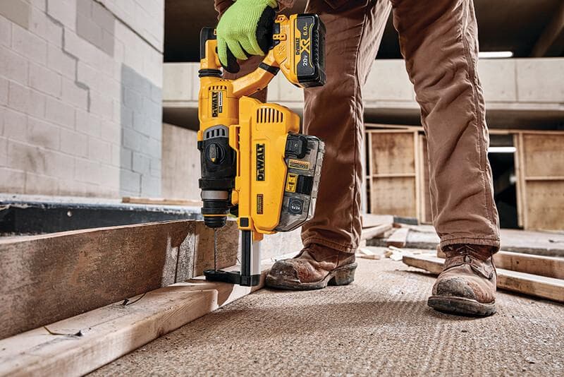 Una persona con pantalones de trabajo marrones, botas de seguridad y un guante verde está usando un martillo rotativo inalámbrico amarillo DEWALT con accesorio de extracción de polvo para perforar un trozo de madera en una obra de construcción.