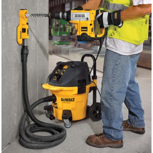 Una persona utiliza una herramienta DEWALT para perforar una pared de concreto, con un accesorio de extracción de polvo DEWALT y un sistema de aspiradora DEWALT conectados para la recolección de polvo. La persona lleva guantes, chaleco de seguridad y botas de trabajo en un entorno industrial.