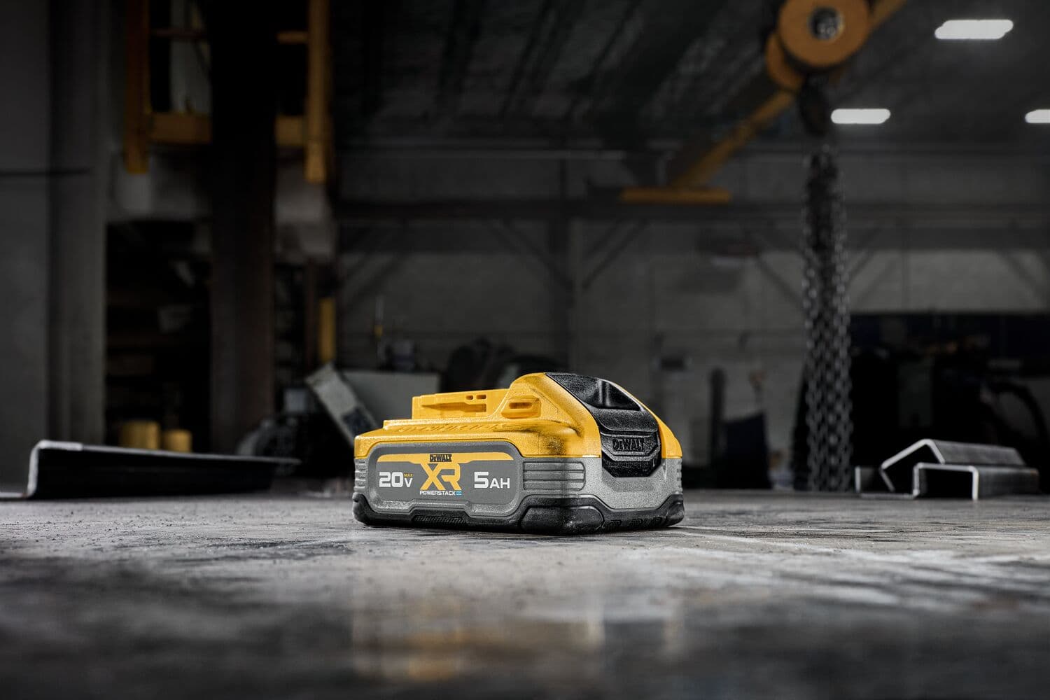 A DEWALT 20V XR 5Ah PowerStack lithium-ion battery resting on a concrete surface in a dimly lit industrial setting. The battery is yellow and black with labeling visible on its side.