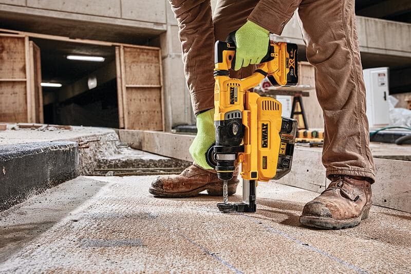 Una persona con guantes verdes y pantalones de trabajo marrones utiliza un martillo rotativo inalámbrico amarillo DEWALT con accesorio de extracción de polvo para perforar un suelo de concreto en una obra de construcción.