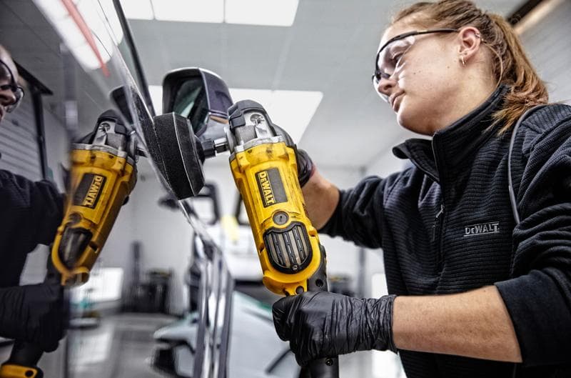 Una persona con guantes negros y una chaqueta DEWALT está usando una pulidora amarilla DEWALT DWP849X en el lateral de un coche, con el reflejo de la herramienta visible en la superficie del coche. El rostro de la persona está difuminado.