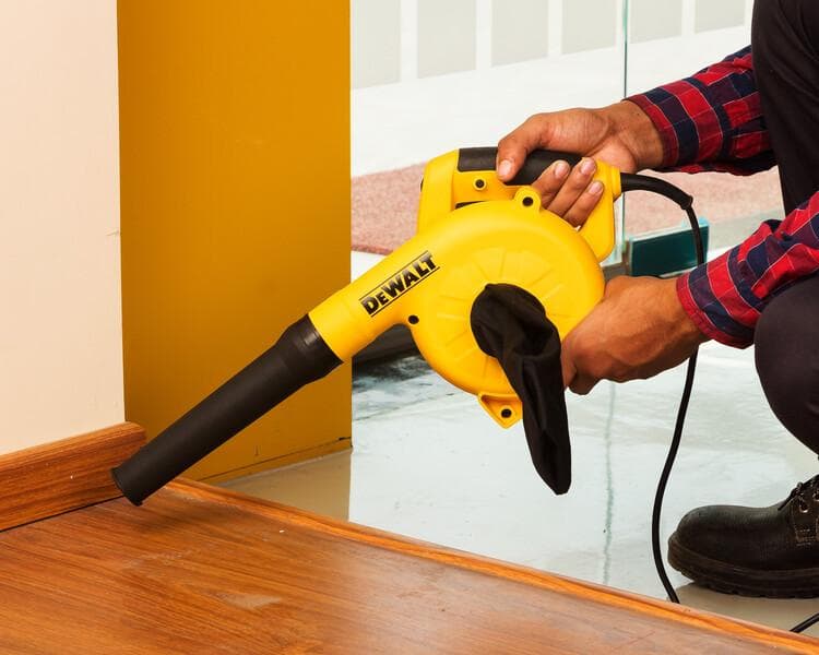 Una persona utiliza un soplador eléctrico amarillo DEWALT para limpiar el polvo de la esquina de un piso de madera cerca de una pared.