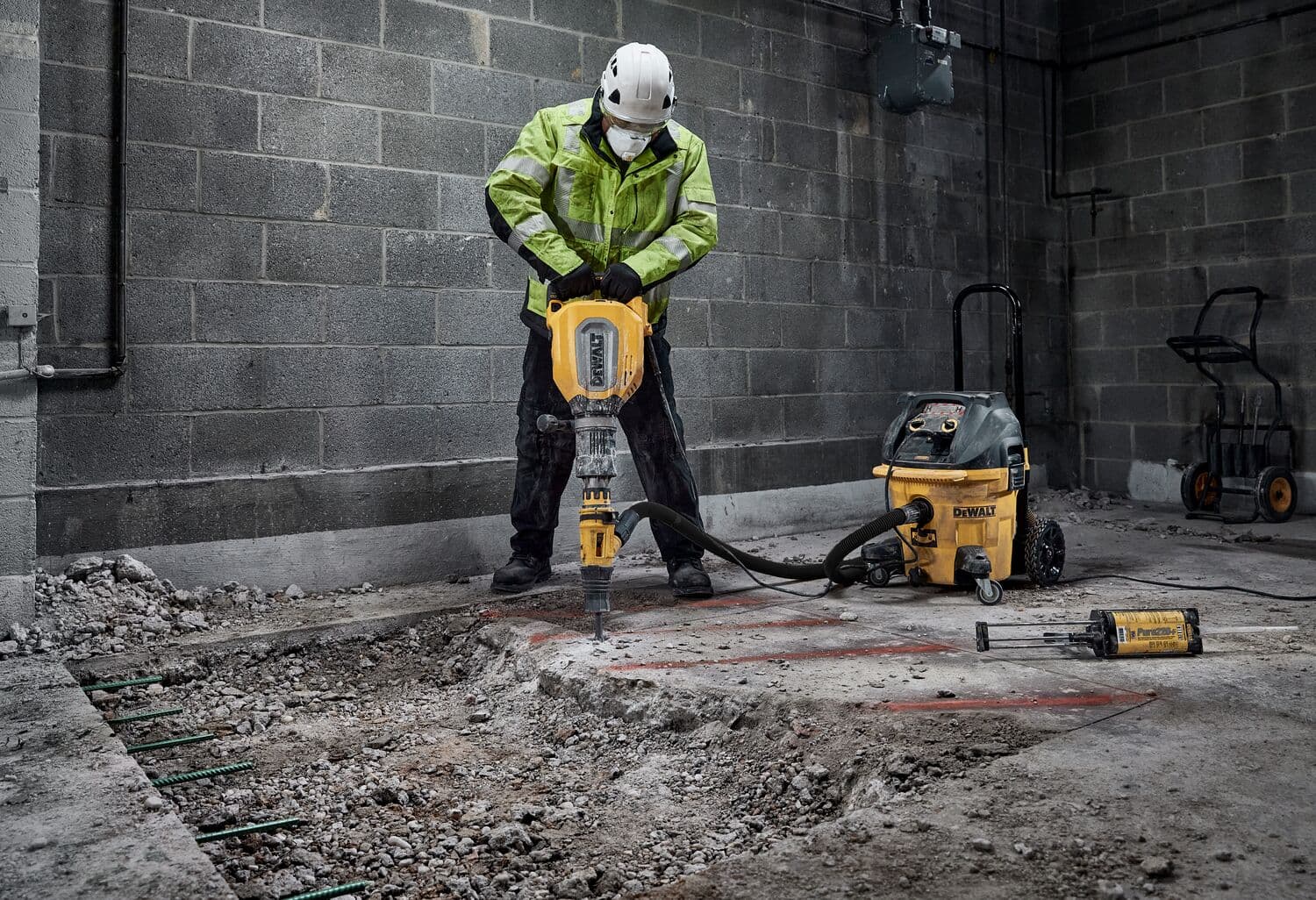 Un trabajador de la construcción con casco y chaqueta de seguridad está utilizando un martillo neumático DEWALT para romper concreto dentro de un edificio industrial. Cerca, hay una aspiradora DEWALT y otras herramientas en el suelo.