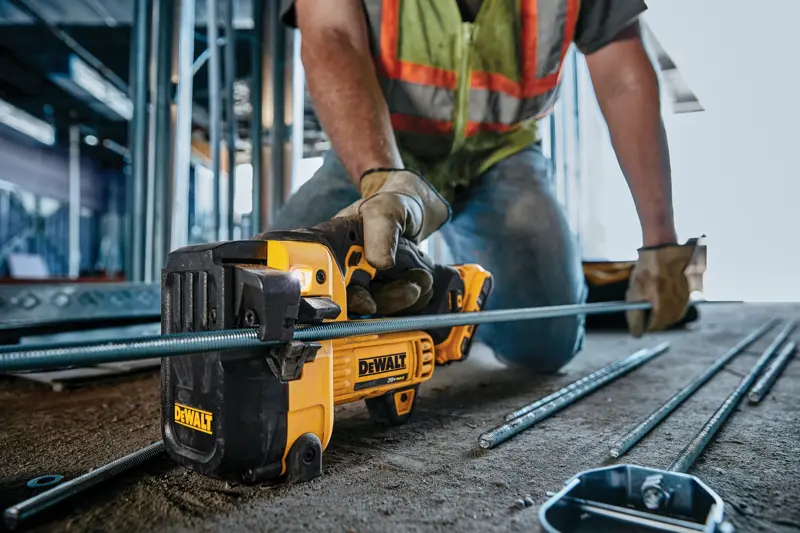 Una persona con guantes de trabajo y chaleco de seguridad usa una herramienta de corte inalámbrica DEWALT para cortar una varilla metálica en una obra de construcción.
