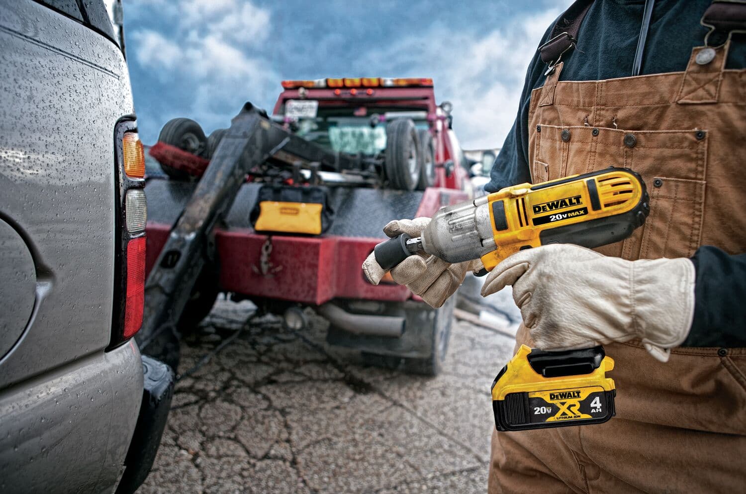 Una persona con un mono marrón y guantes sostiene una llave de impacto inalámbrica DEWALT 20V MAX cerca de un vehículo y una grúa roja. El entorno parece ser al aire libre, posiblemente junto a una carretera, y la herramienta DEWALT está claramente en uso.