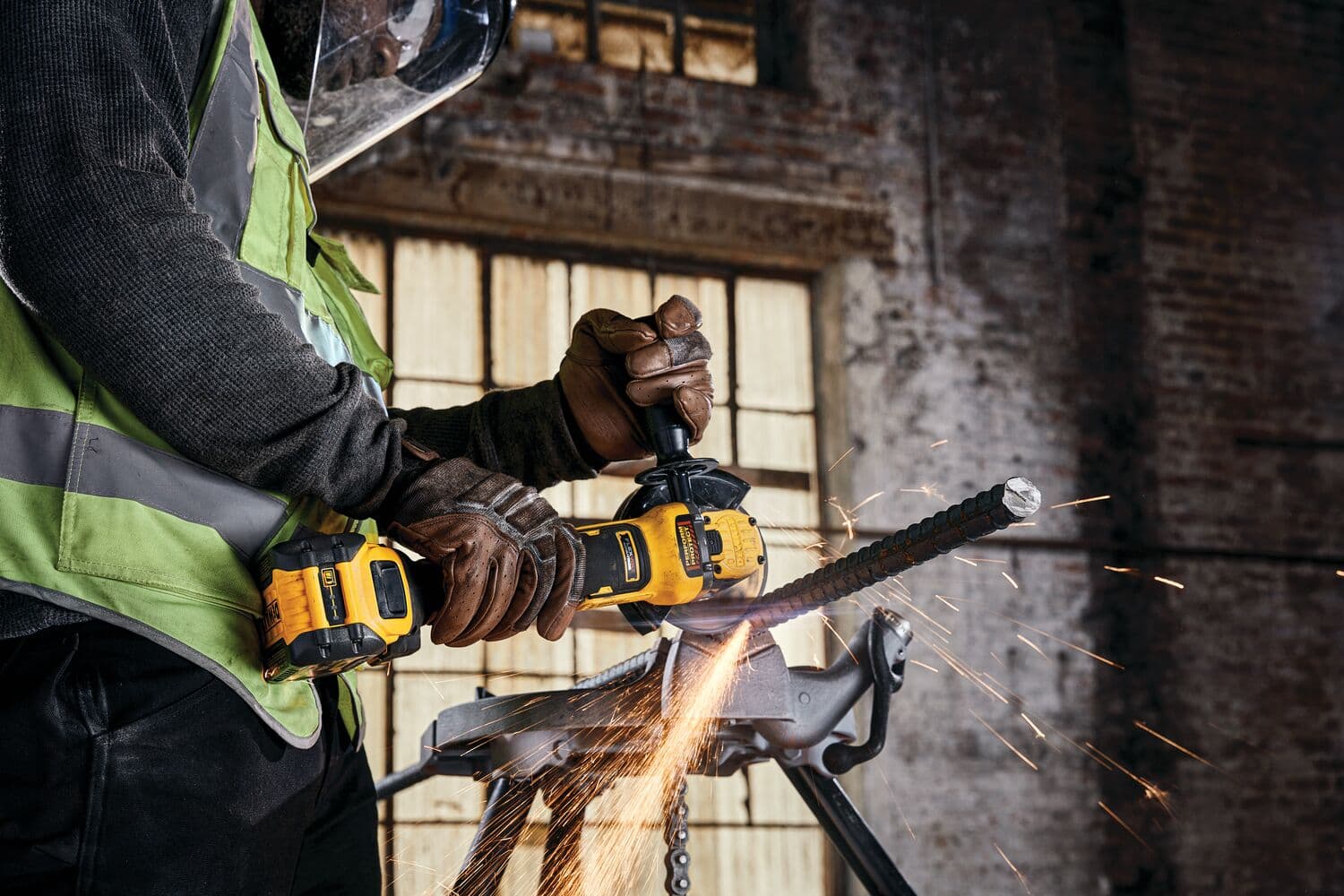 Una persona con guantes protectores y un chaleco de seguridad utiliza una amoladora angular inalámbrica DEWALT para cortar una barra de metal, generando chispas. El entorno parece ser un espacio industrial con grandes ventanas y paredes de ladrillo expuesto.