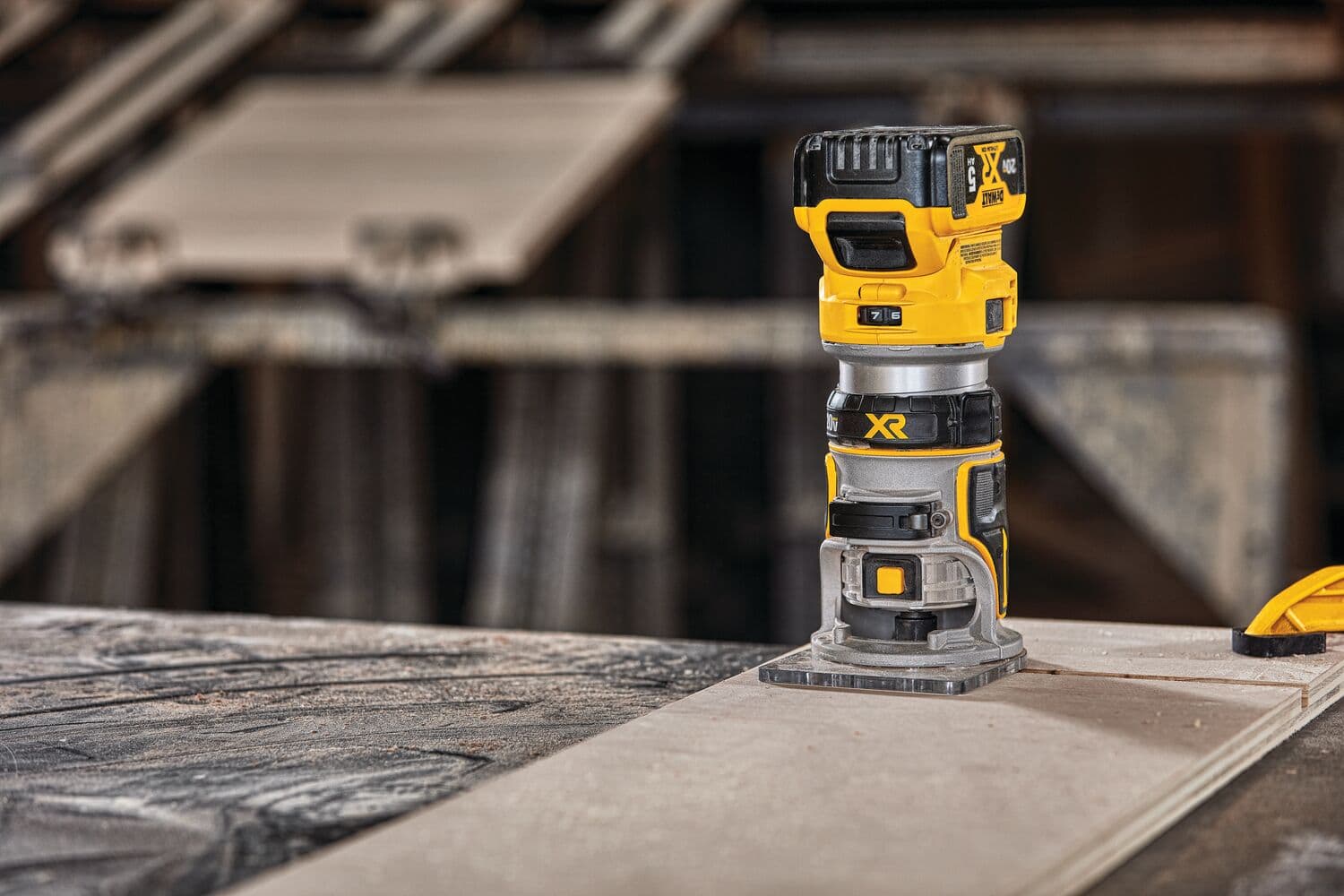A DEWALT cordless compact router, model DCW600B, is placed upright on a wooden work surface in a workshop setting. The tool features a yellow and black design and is used for woodworking.