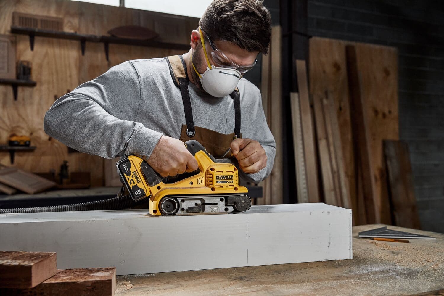 Una persona usa una lijadora de banda inalámbrica DEWALT DCW220B para suavizar un trozo de madera en un taller. El rostro de la persona está borroso por privacidad. Se pueden ver varias piezas de madera y herramientas en el fondo.