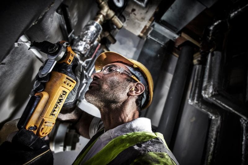 Una persona con casco de seguridad amarillo y chaleco utiliza una herramienta eléctrica DEWALT DWE315 para cortar o lijar material en una pared en un entorno industrial. Su rostro está oculto.