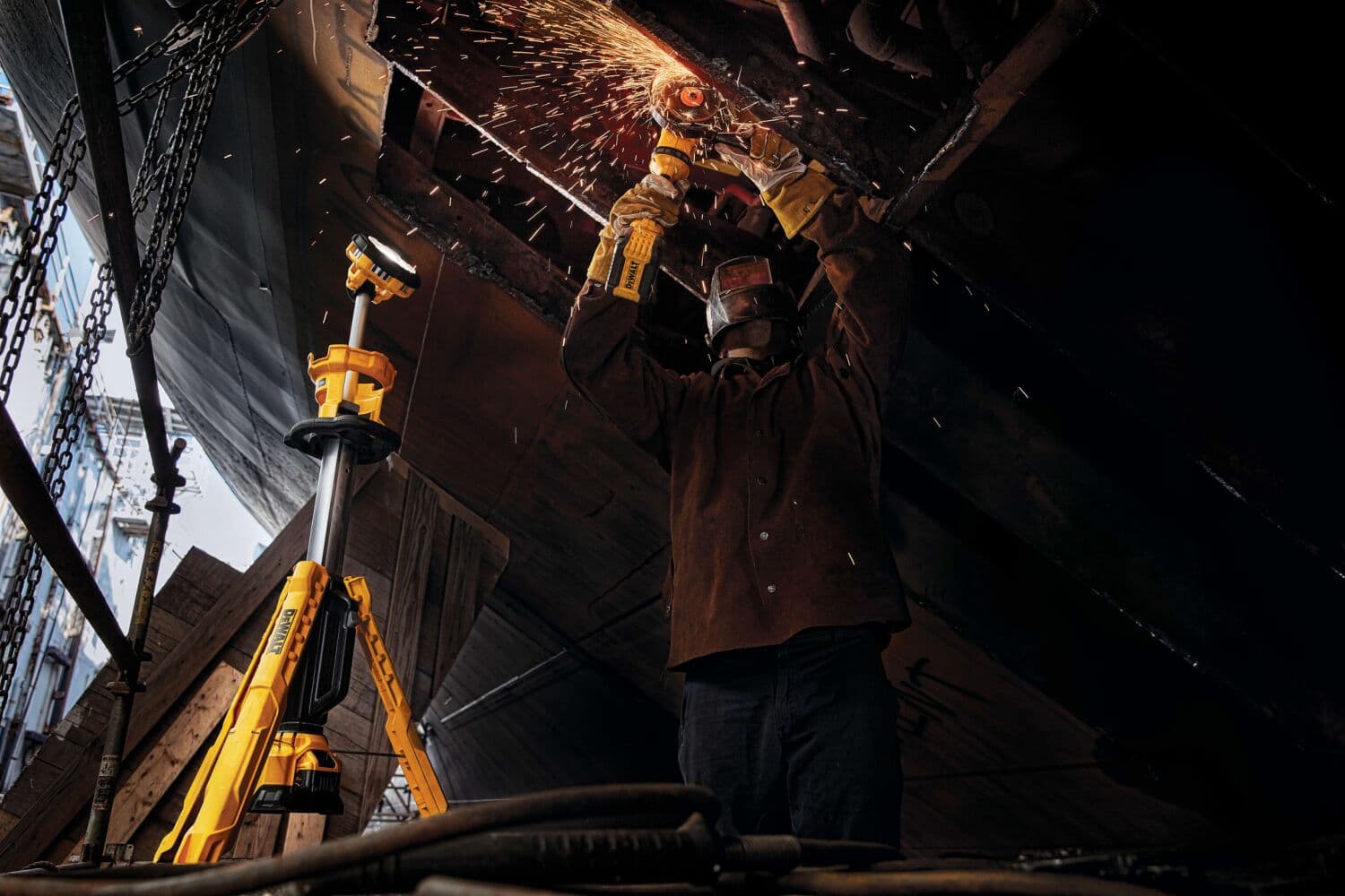 Un trabajador con equipo de protección utiliza una herramienta eléctrica para pulir metal, mientras salen chispas. Junto al trabajador hay una luz de trabajo amarilla DEWALT con trípode, posicionada para iluminar el área de trabajo.