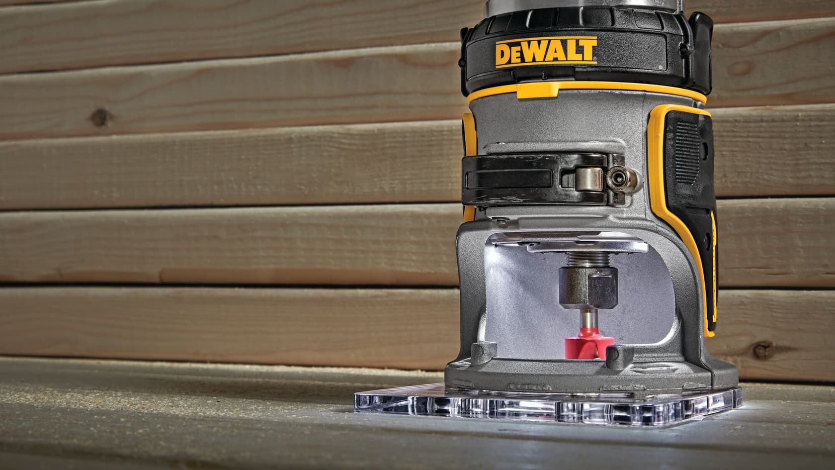 Imagen en primer plano de una fresadora inalámbrica DEWALT colocada sobre una superficie de madera, con paneles de madera en el fondo. La herramienta se muestra en uso con una fresa instalada y luz LED iluminando el área de trabajo.