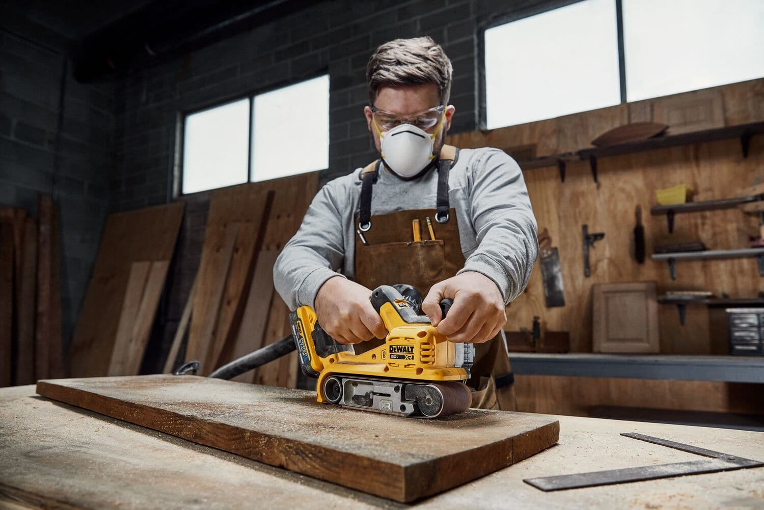 Una persona utiliza una lijadora de banda inalámbrica DEWALT DCW220B para lijar una pieza de madera en un banco de trabajo en un taller.