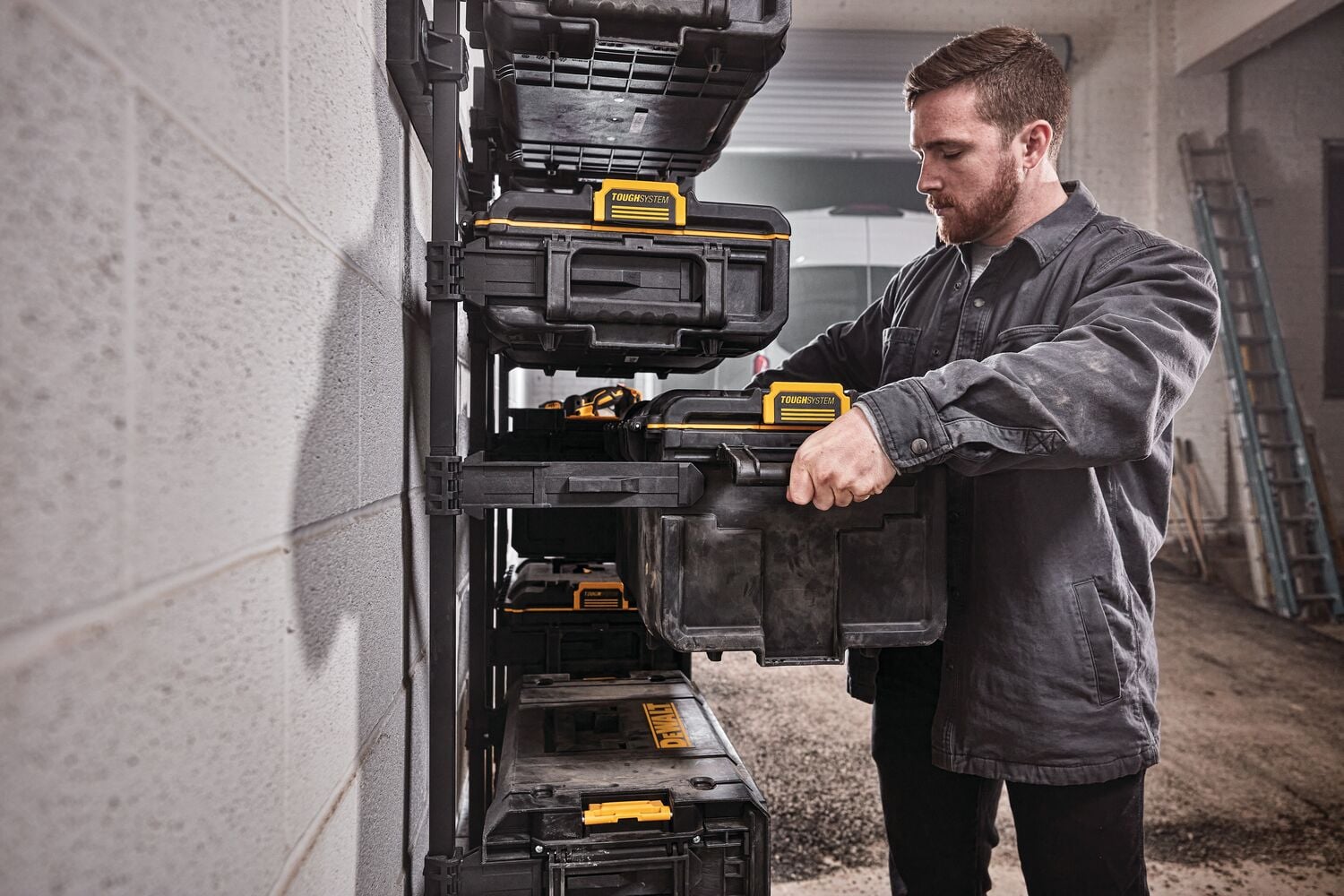 Una persona está organizando cajas de almacenamiento de herramientas DEWALT negras y amarillas en una estantería montada en la pared dentro de un taller.