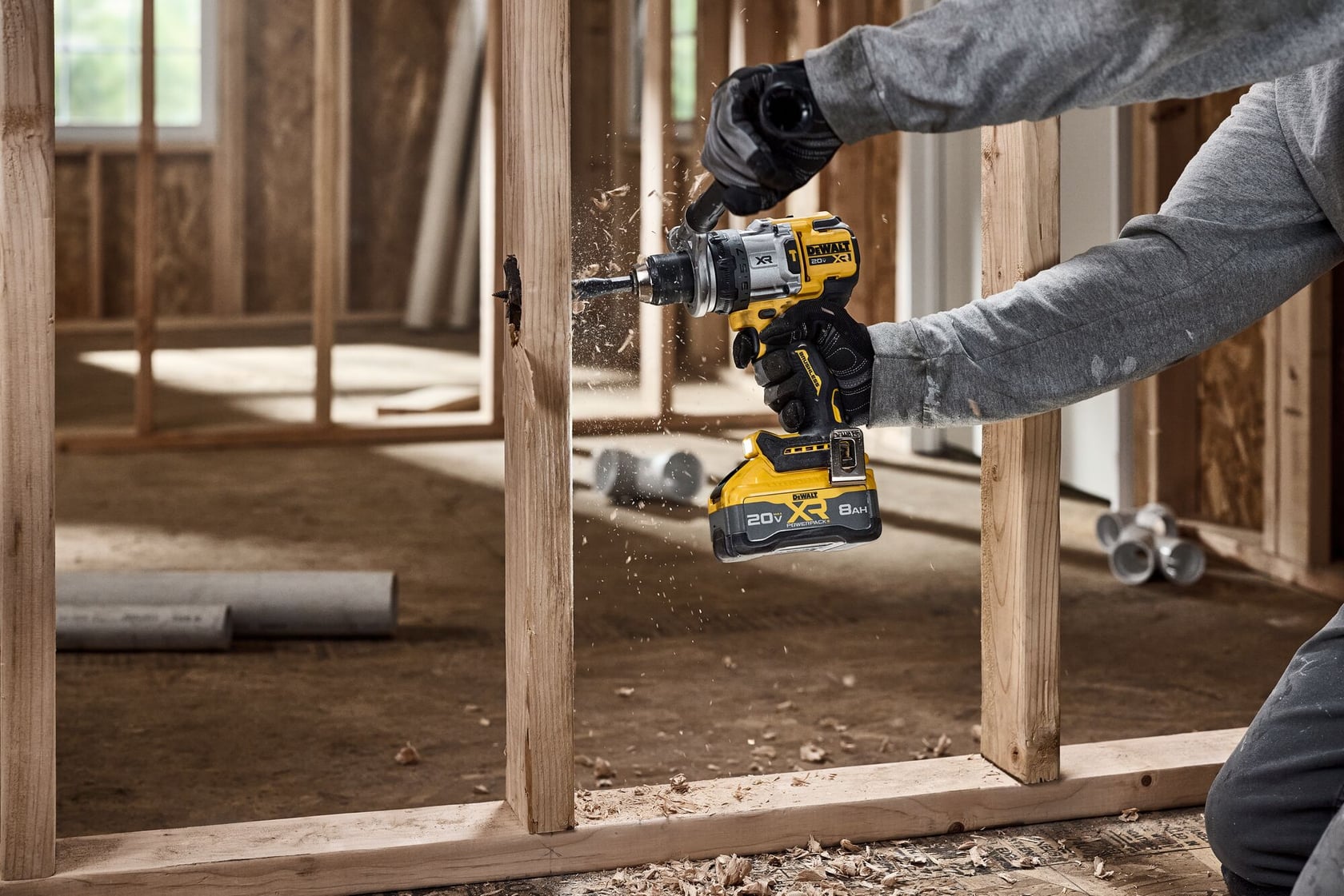 A person is using a DEWALT cordless drill to make a hole in a wooden stud inside a construction site. Wood shavings are visible as the drill operates, and the person is wearing gloves and a gray long-sleeve shirt.