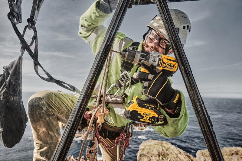 Un trabajador con equipo de seguridad y guantes utiliza un taladro inalámbrico DEWALT DCD996P2 al aire libre, realizando tareas de mantenimiento o instalación sobre una estructura metálica encima de un terreno rocoso cerca del mar.