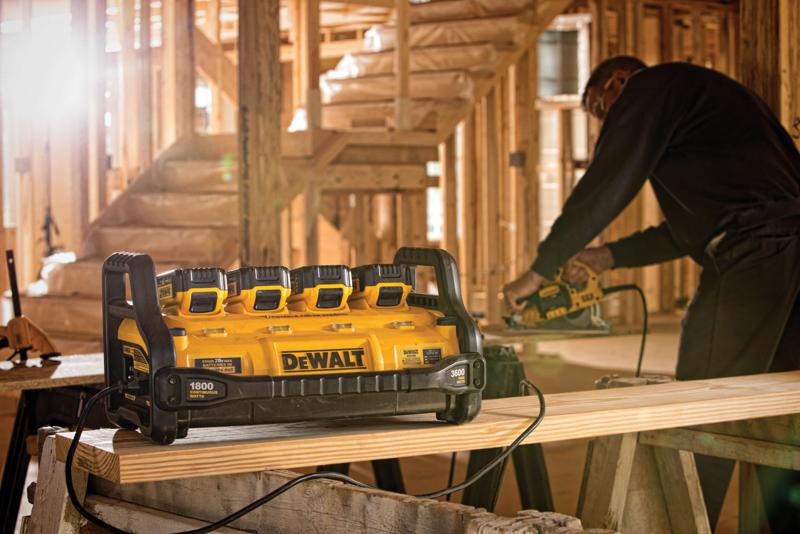 Una estación de energía portátil DEWALT modelo DCB1800B con cuatro ranuras para baterías está colocada sobre una tabla de madera en un edificio en construcción, con una persona usando una herramienta eléctrica al fondo.