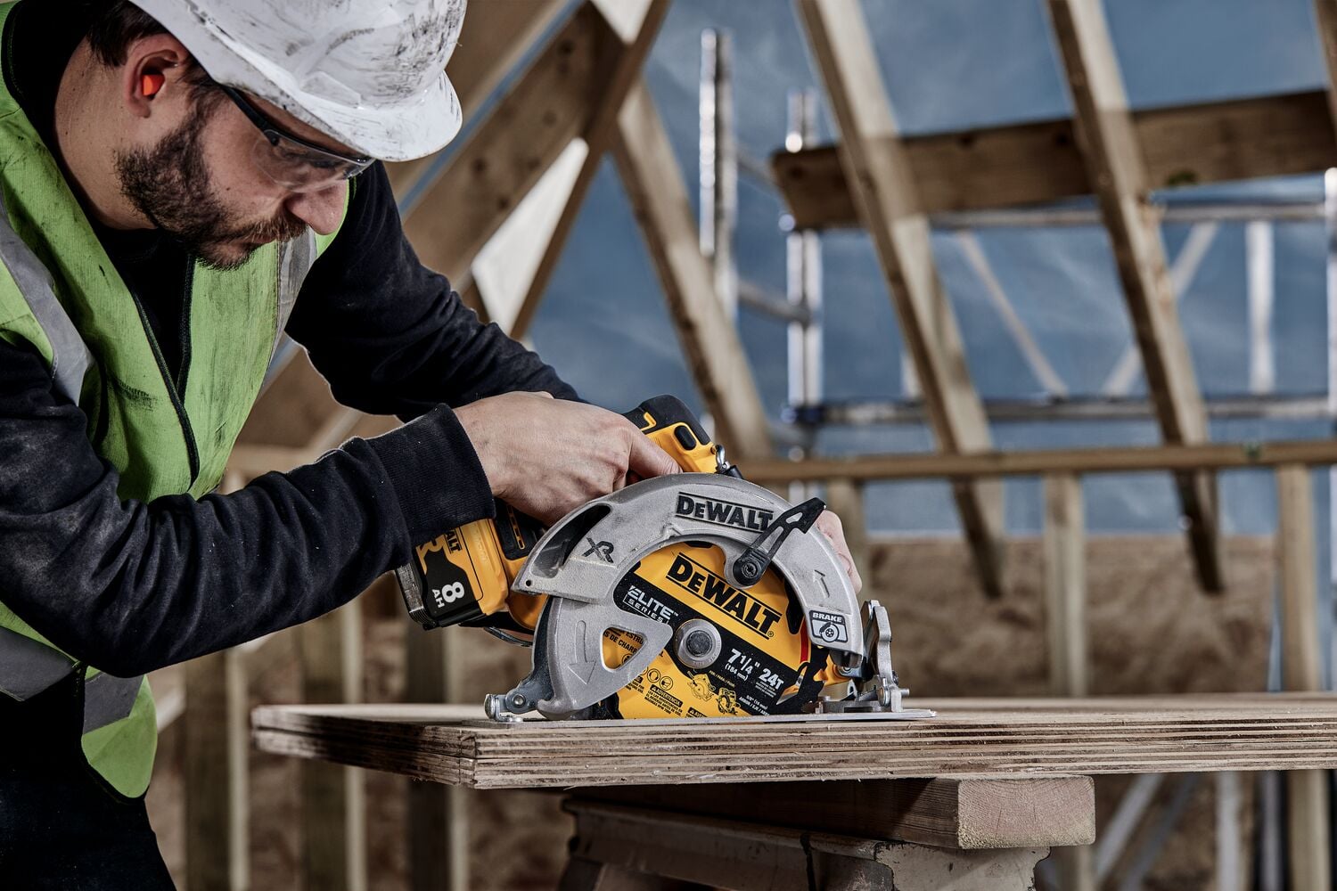 Una persona con casco de seguridad y chaleco está usando una sierra circular DEWALT para cortar tablas de madera en un sitio de construcción con vigas de madera expuestas al fondo.