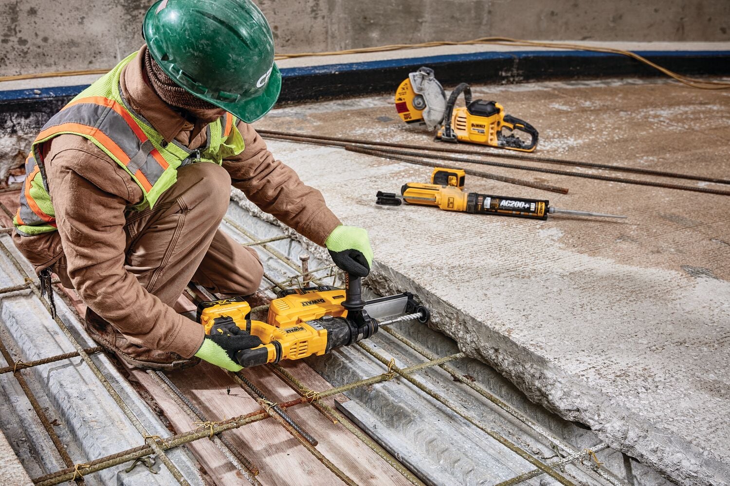 Un trabajador de la construcción con equipo de seguridad utiliza una herramienta eléctrica amarilla DEWALT para perforar concreto en una obra. Otras herramientas DEWALT están visibles en el suelo cerca de él.