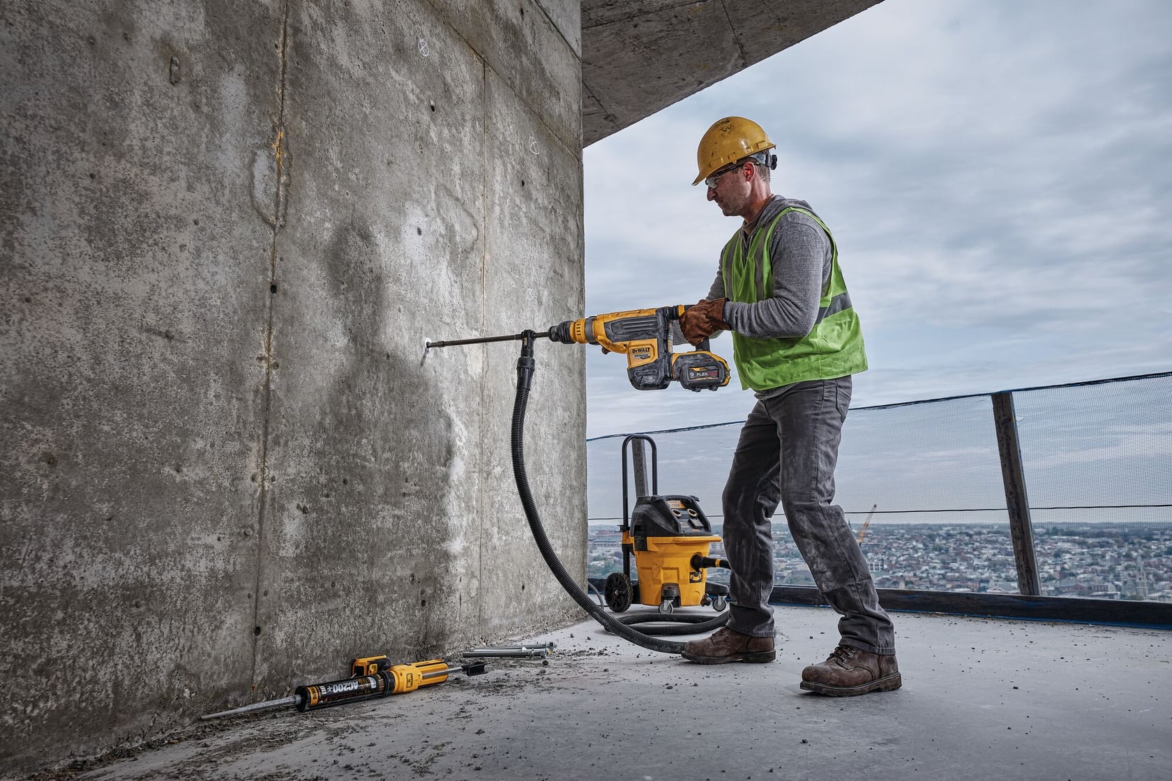 Un trabajador de construcción con un casco amarillo y un chaleco de seguridad verde utiliza un taladro rotativo inalámbrico DEWALT para perforar una pared de concreto. La herramienta está conectada a un extractor de polvo a través de una manguera, y otros equipos DEWALT se ven en el suelo cerca.
