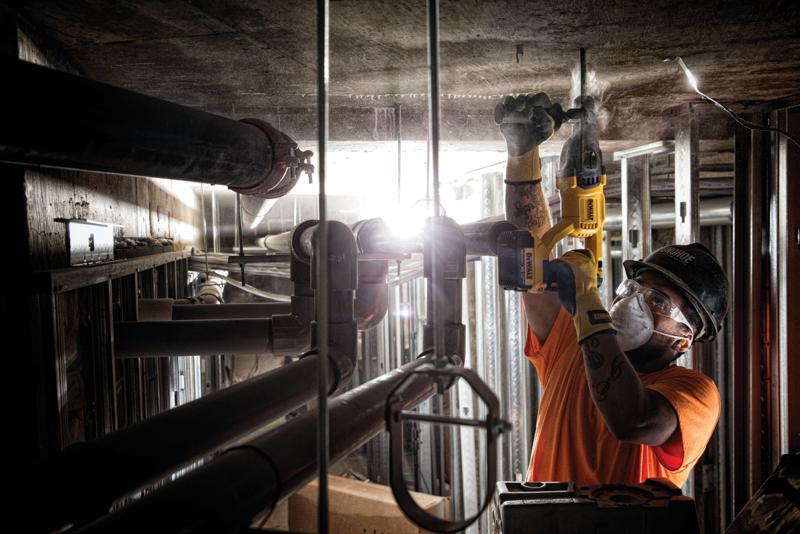 Una persona con casco y guantes de seguridad utiliza un martillo perforador amarillo DEWALT para trabajar en un techo de concreto dentro de un sitio de construcción industrial. Se pueden ver tuberías y estructuras metálicas alrededor del trabajador.