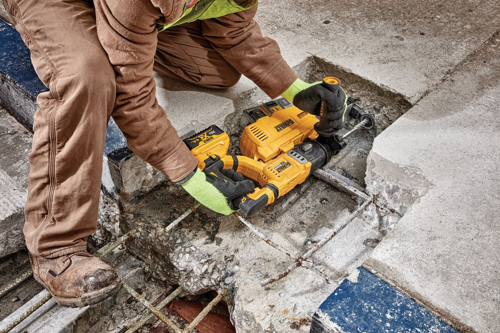 Una persona con guantes de trabajo y ropa de trabajo marrón utiliza un martillo perforador inalámbrico amarillo DEWALT para romper el hormigón y exponer barras de metal en un sitio de construcción.