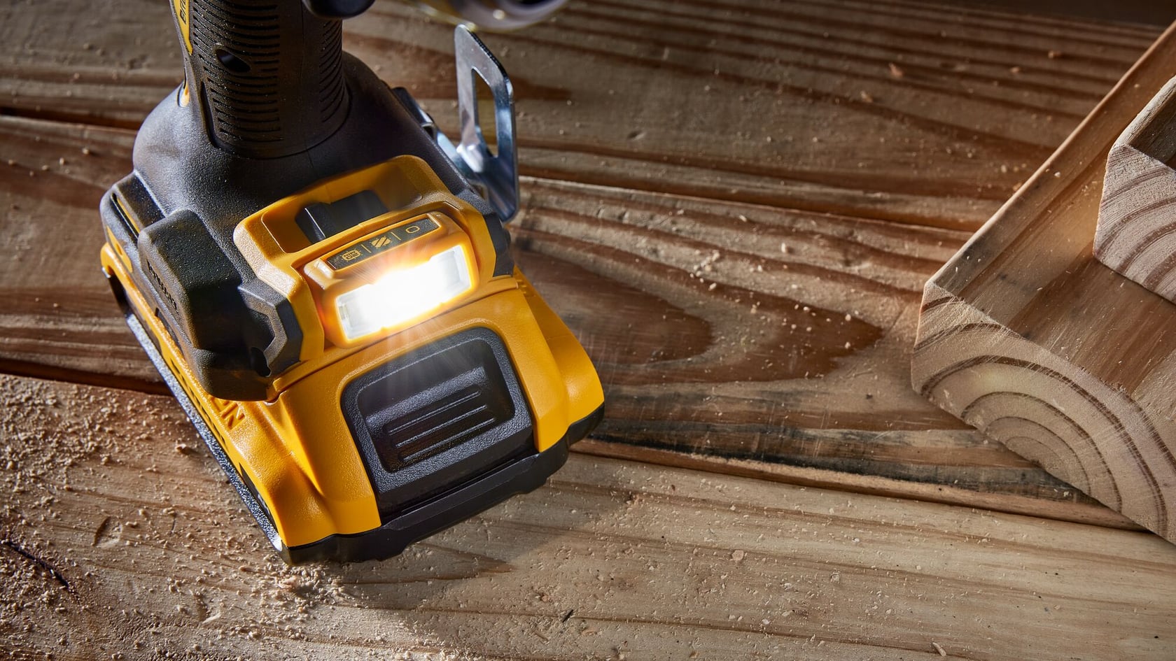 Vista de cerca de la base de una herramienta eléctrica inalámbrica amarilla y negra con una luz LED encendida, colocada sobre una superficie de madera con aserrín y una pieza de madera cerca.