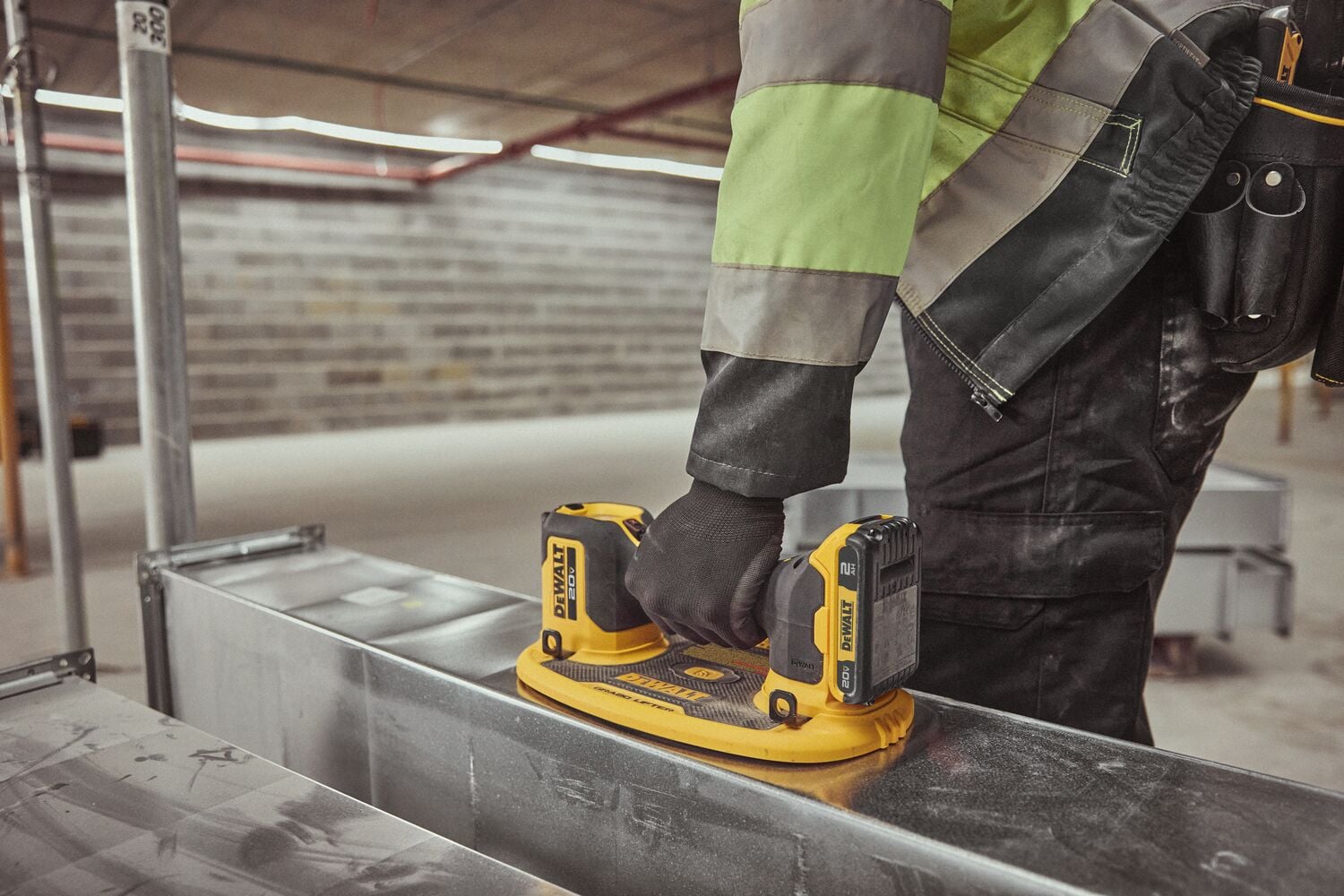 A person wearing work gloves and a high-visibility jacket operates a DEWALT cordless power tool on a metal surface in an industrial setting.