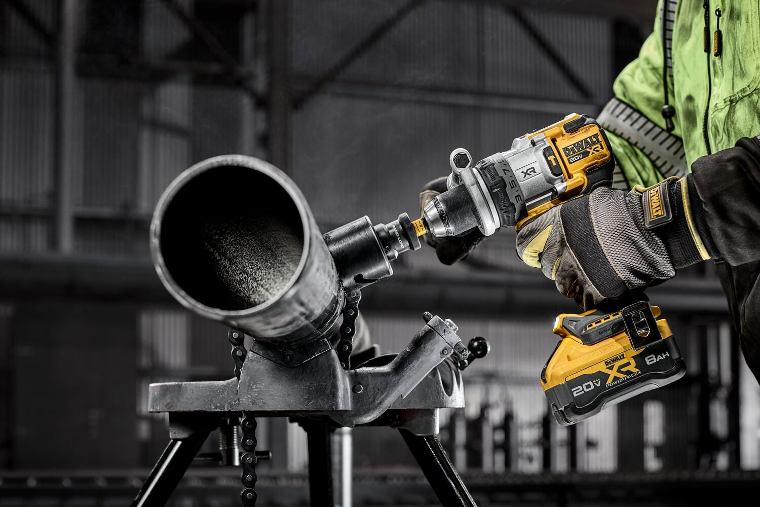 A person wearing safety gear is using a DEWALT cordless power drill to cut or drill into a large metal pipe, which is secured on a stand in an industrial setting.