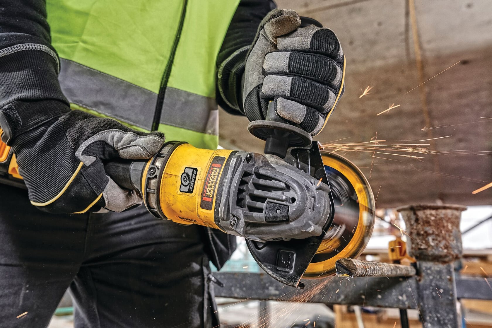 Primer plano de una persona con guantes de seguridad y chaleco reflectante, usando una amoladora angular amarilla para cortar un tubo de metal. Se pueden ver chispas en la zona de corte.