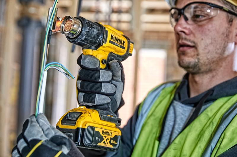 Una persona con guantes y chaleco de seguridad está usando una pistola de calor inalámbrica DEWALT 18V XR para trabajar en cables eléctricos en un sitio de construcción. El rostro de la persona está difuminado.