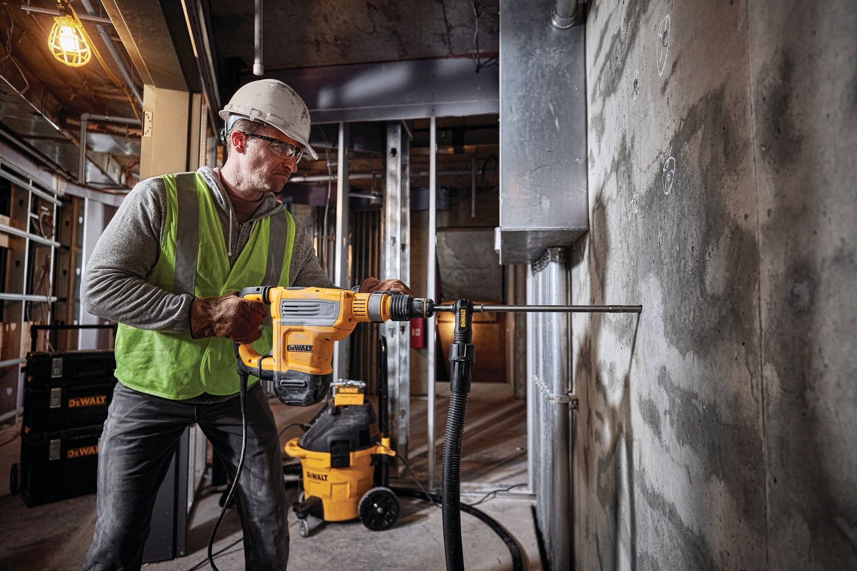 Un trabajador con casco de seguridad y chaleco opera un taladro rotativo DEWALT (SKU: D25614K) para perforar una pared de concreto en un sitio de construcción. Se pueden ver herramientas y equipos DEWALT en el fondo.