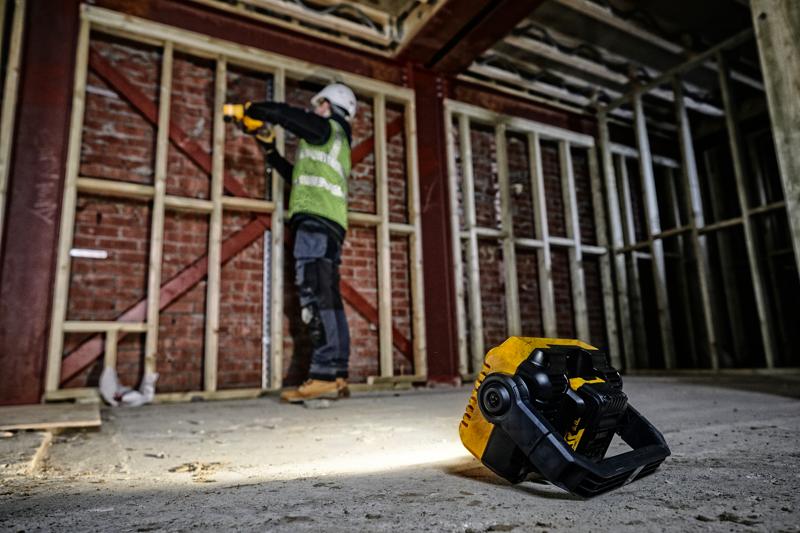 Una luz de trabajo portátil DEWALT (SKU: DCL077) está colocada en el suelo de una obra, iluminando el área. Al fondo, un trabajador de la construcción con equipo de seguridad trabaja cerca de una estructura de madera junto a una pared de ladrillo.