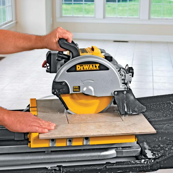 Una sierra para azulejos DEWALT D24000 está siendo utilizada para cortar una baldosa de cerámica en una habitación luminosa con suelo de baldosas. Una persona sostiene el mango de la sierra y guía la baldosa para un corte preciso. Se observa agua, lo que indica el uso de un método de corte húmedo.