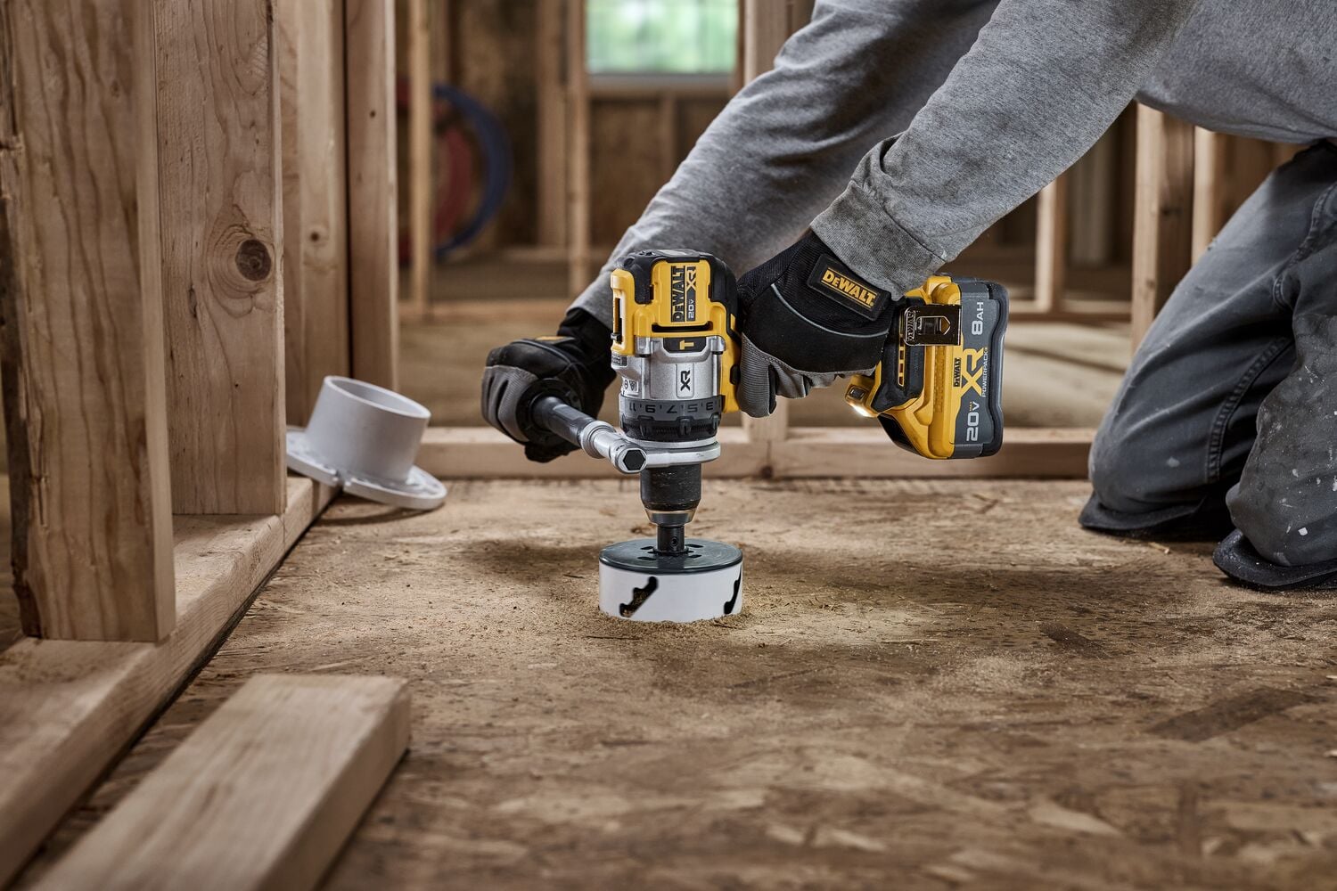 A person is using a DEWALT cordless power drill with a hole saw attachment to cut into a wooden subfloor inside a construction site with exposed wooden framing.