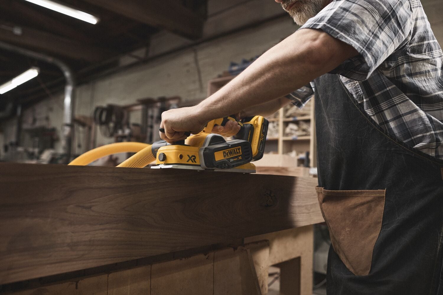 Una persona usa una sierra de carril inalámbrica DEWALT (modelo DCS520B) para cortar una pieza de madera en un taller de carpintería. La persona lleva un delantal y una camisa de cuadros.