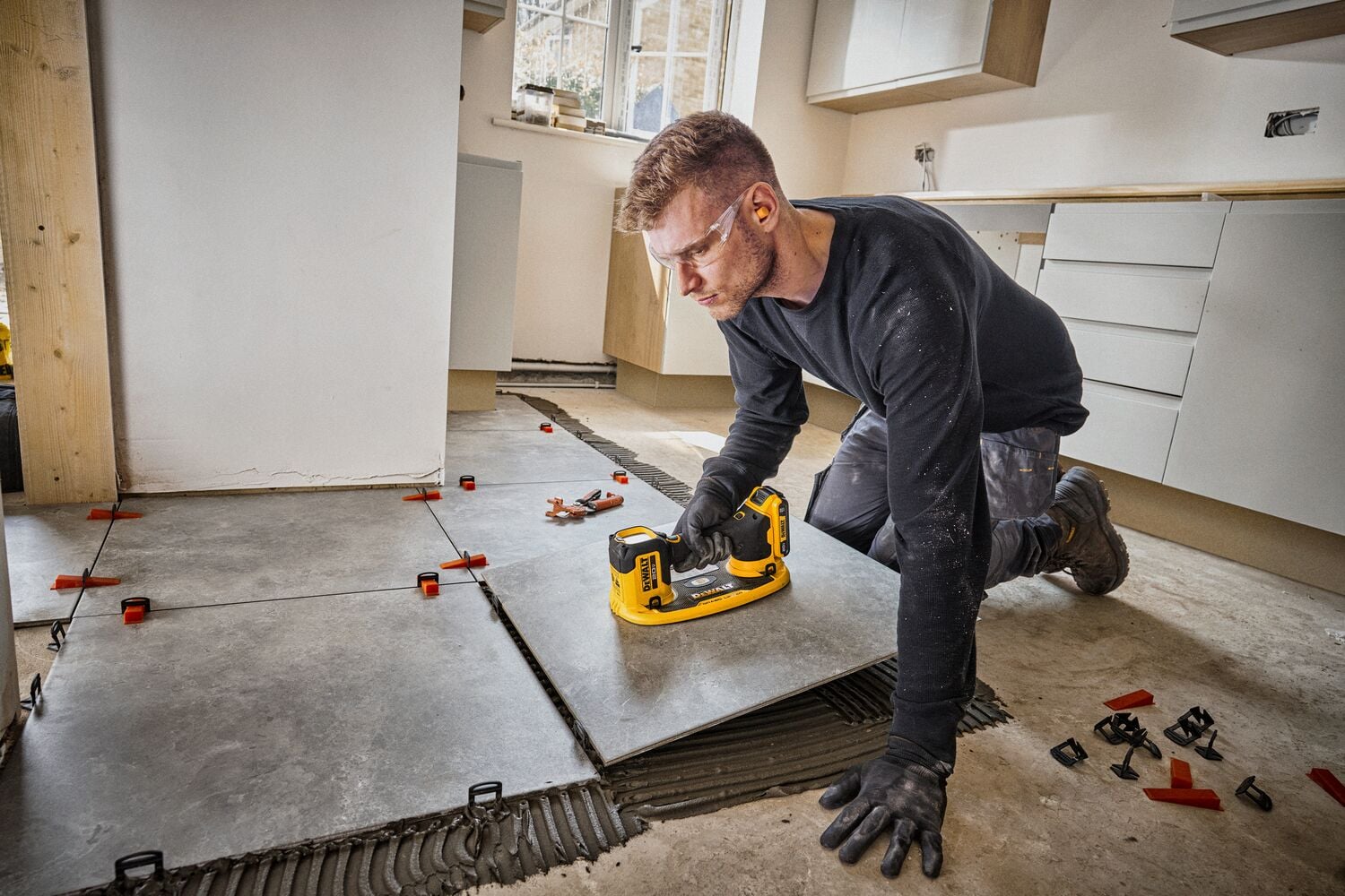 Una persona arrodillada en una cocina está usando una herramienta eléctrica amarilla DEWALT para instalar baldosas grandes de color gris en el suelo. Se ven separadores entre las baldosas y el rostro de la persona está difuminado por privacidad.