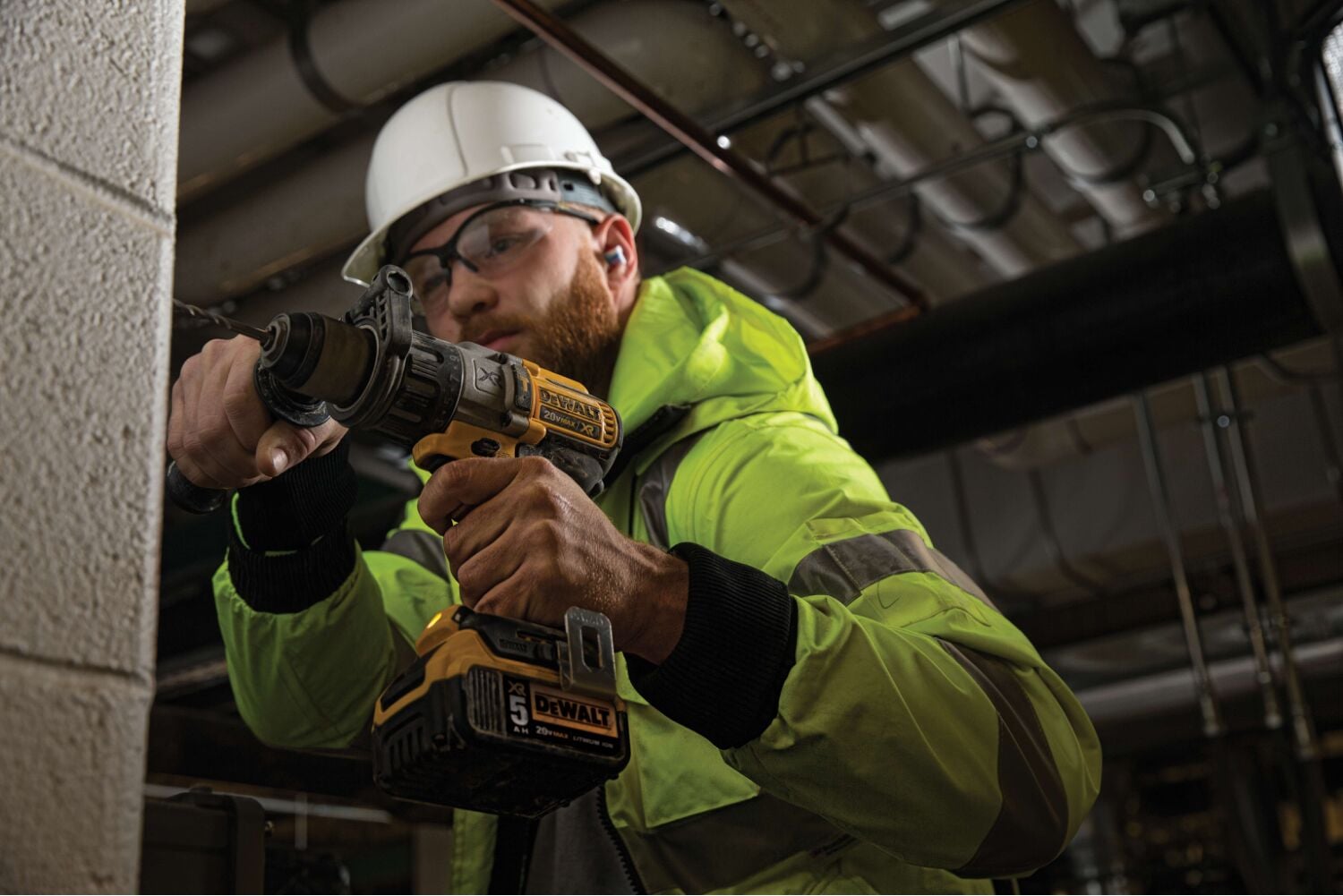 Una persona con chaqueta de seguridad reflectante y casco blanco utiliza un taladro inalámbrico DEWALT amarillo y negro para trabajar en una pared de concreto en un entorno industrial.