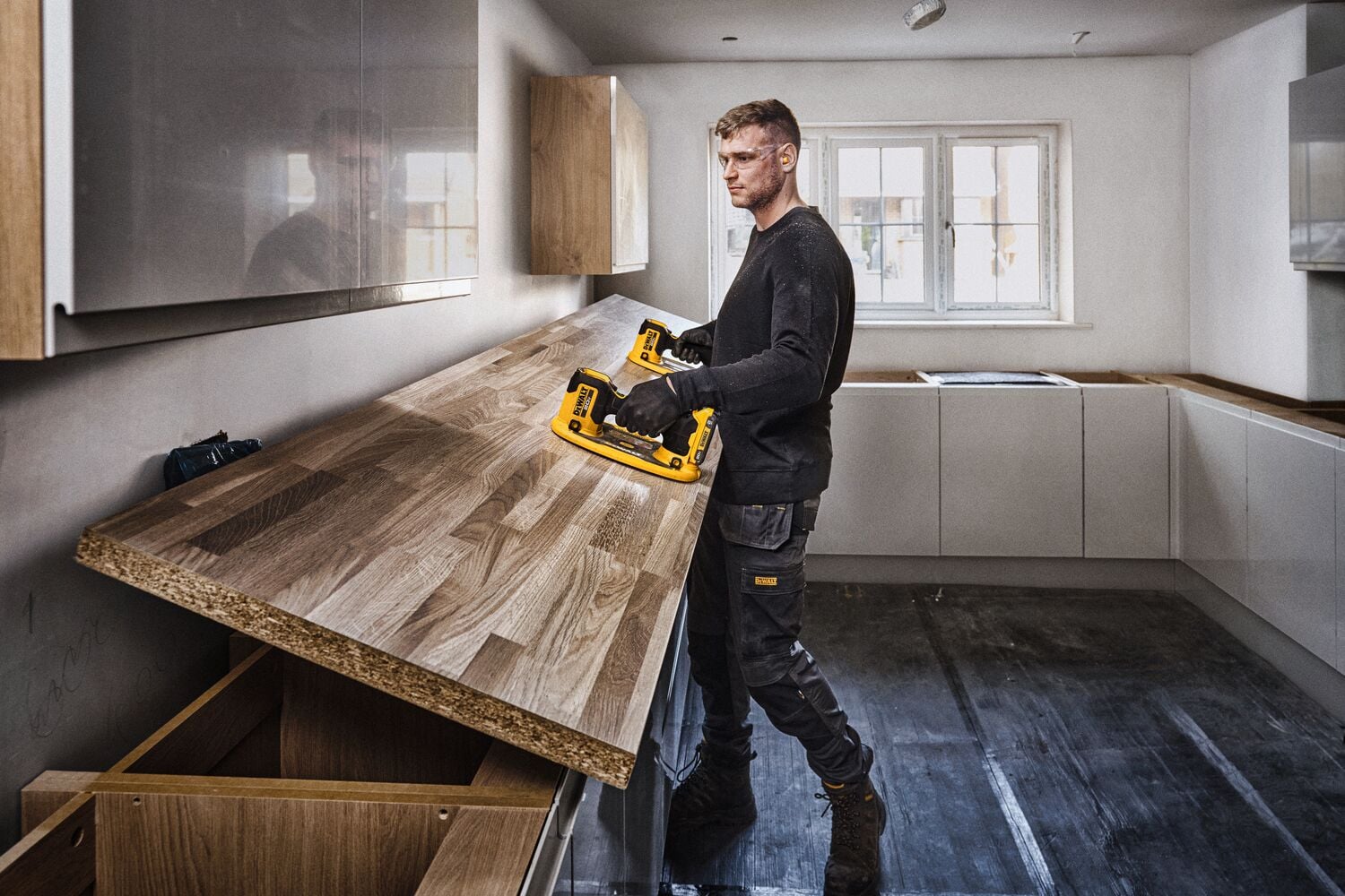 Una persona con el rostro difuminado está instalando una encimera de madera en una cocina usando una herramienta eléctrica DEWALT. La cocina es moderna, con gabinetes de colores claros y una ventana grande en el fondo.