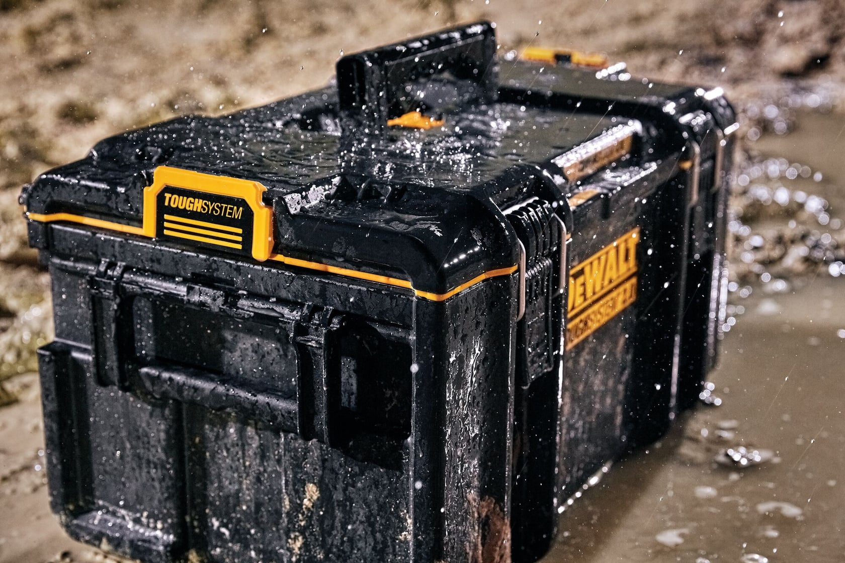 Una caja de herramientas DEWALT ToughSystem negra y amarilla sobre suelo mojado y lodoso, siendo salpicada por agua, demostrando su durabilidad y diseño impermeable.