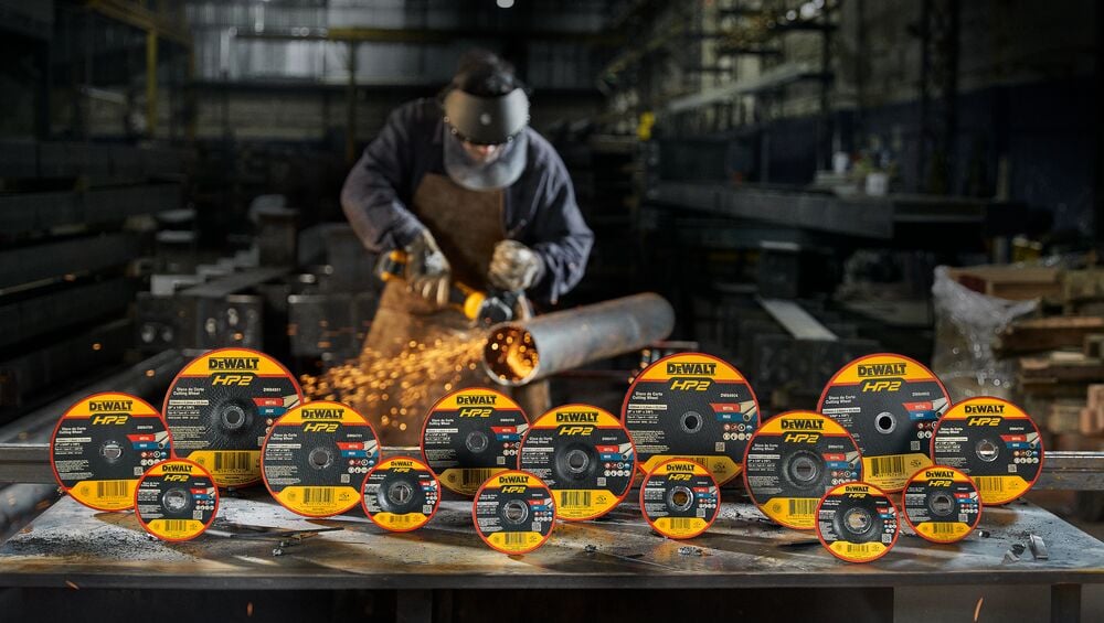A group of DEWALT HP2 abrasive cutting and grinding discs are displayed in the foreground, arranged on a workshop table. In the background, a worker wearing protective gear uses a DEWALT grinder on a metal pipe, producing sparks in an industrial setting.