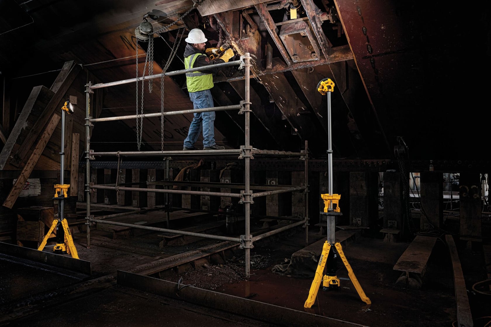 Un trabajador con casco y chaleco de seguridad está de pie en un andamio en un entorno industrial. Hay dos focos de trabajo con trípode amarillos DEWALT iluminando el área.