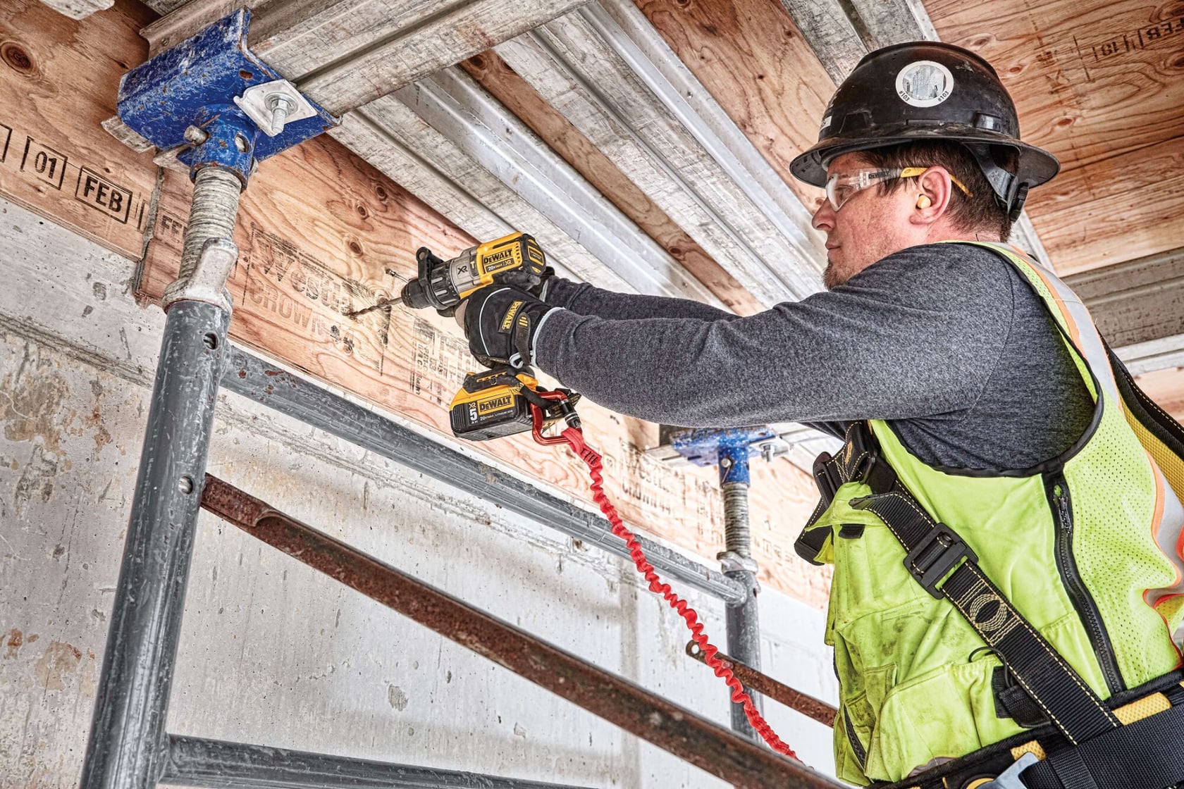 Un trabajador de construcción con casco, chaleco de seguridad y arnés utiliza un taladro inalámbrico para asegurar vigas de metal a una estructura de madera en un sitio de construcción. El trabajador está de pie sobre un andamio y concentrado en su tarea.