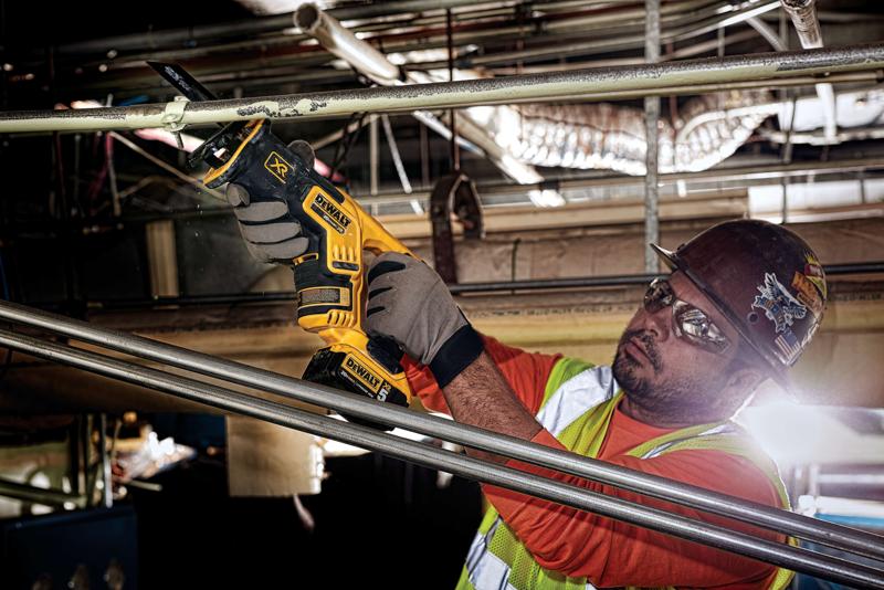 Una persona con guantes de trabajo, camiseta naranja, chaleco de seguridad y casco está usando una sierra recíproca inalámbrica DEWALT DCS367B para cortar una tubería metálica en un entorno industrial. El rostro de la persona está difuminado por privacidad.