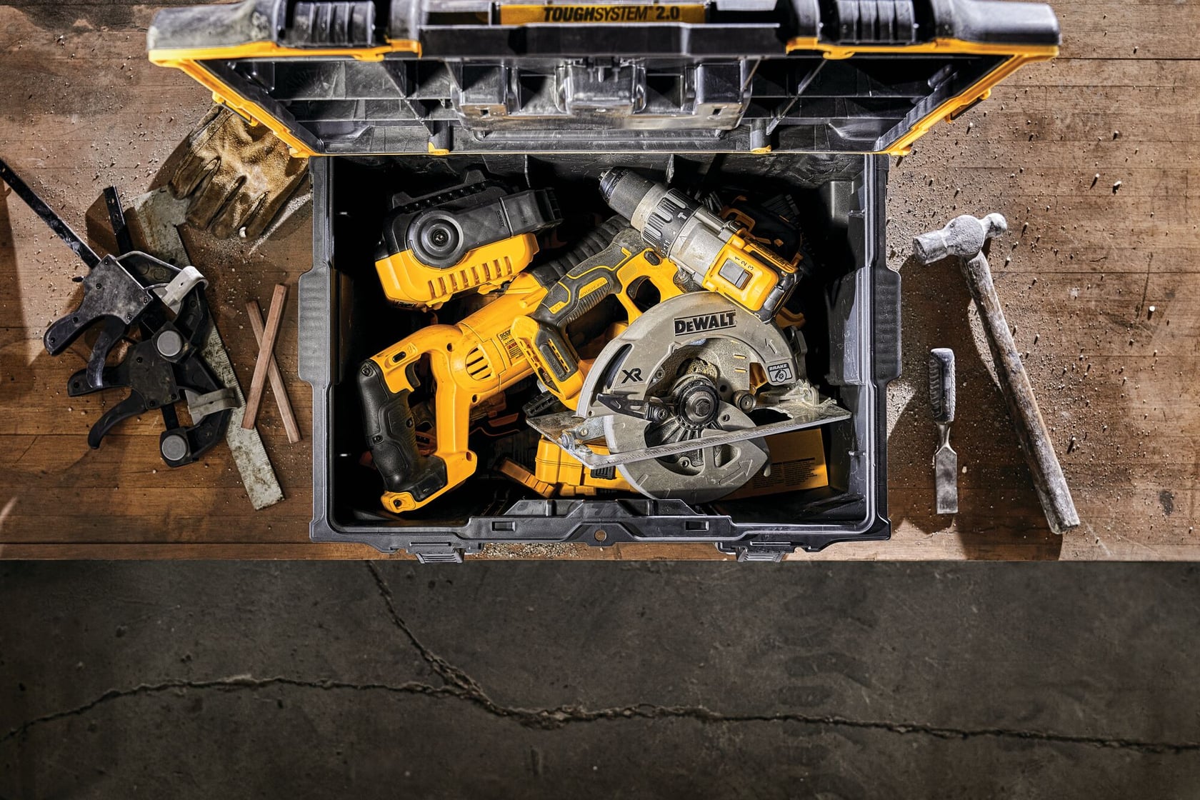 Una caja de herramientas DEWALT ToughSystem 2.0 sobre un banco de trabajo, abierta para mostrar varias herramientas eléctricas amarillas y negras, incluyendo una sierra circular y un taladro. Hay otras herramientas manuales y accesorios dispersos alrededor del banco.