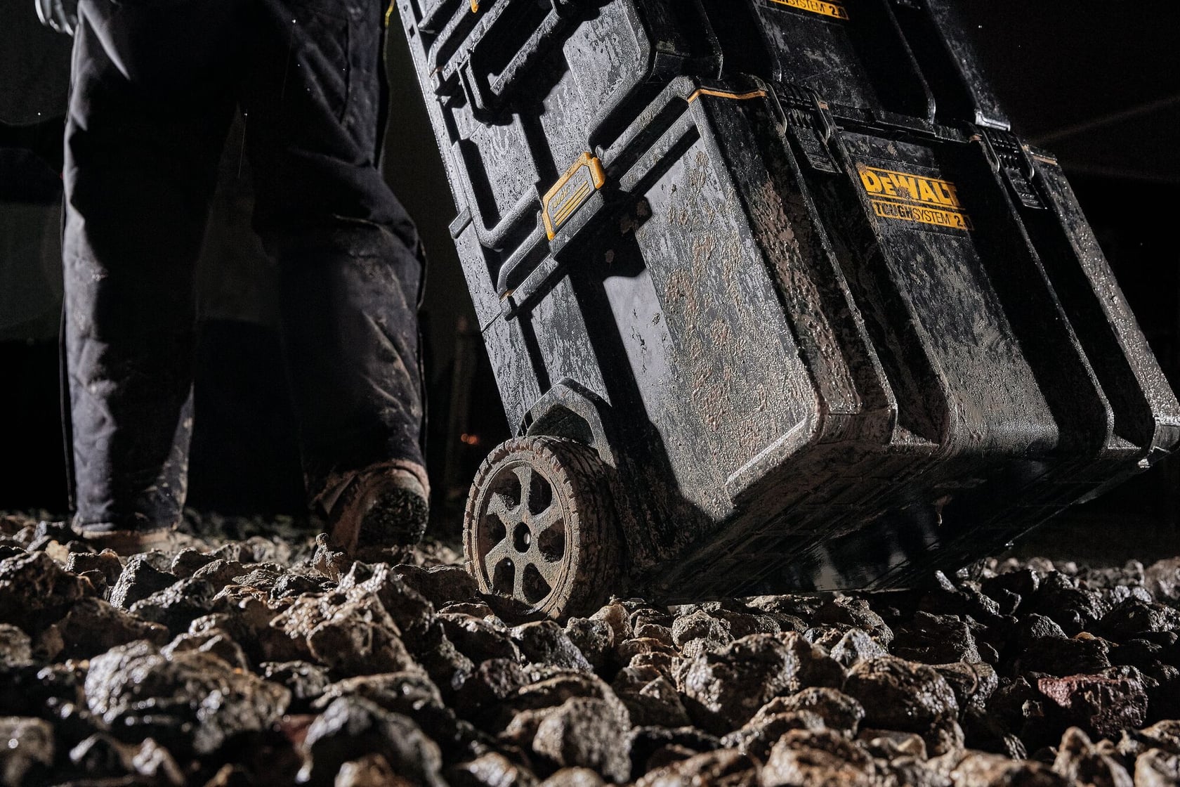 Vista de primer plano de una caja de herramientas con ruedas DEWALT ToughSystem siendo arrastrada sobre terreno rocoso. La caja y las ruedas están cubiertas de barro, y una persona con pantalones de trabajo sucios y botas está de pie cerca.