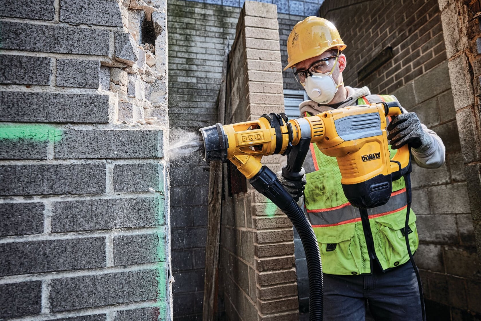Un trabajador de la construcción con casco y chaleco de seguridad utiliza un martillo demoledor amarillo DEWALT D25614K con extracción de polvo sobre una pared de ladrillo.