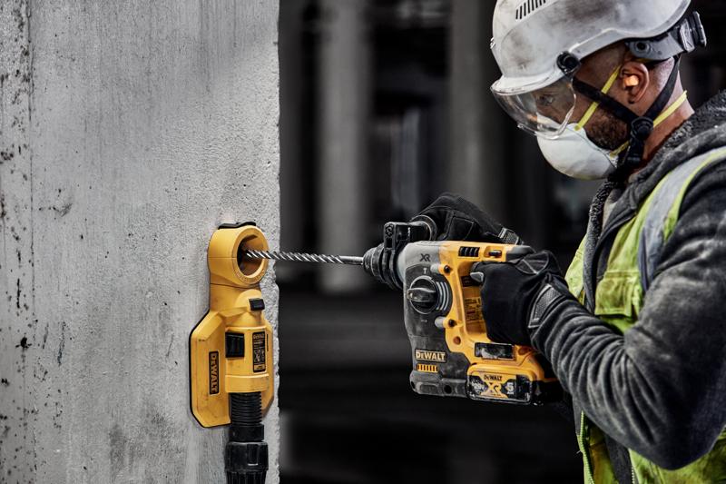 Un trabajador de la construcción con equipo de seguridad utiliza un taladro percutor inalámbrico DEWALT para perforar una pared de concreto. Un accesorio de extracción de polvo DEWALT está conectado a la pared alrededor de la broca para controlar el polvo.