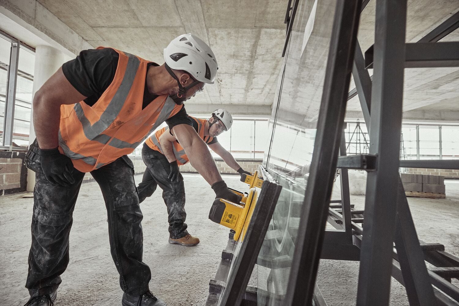 Two workers in safety helmets and orange vests use a yellow DEWALT tool to assist with the installation or movement of a large sheet of glass in a construction site.