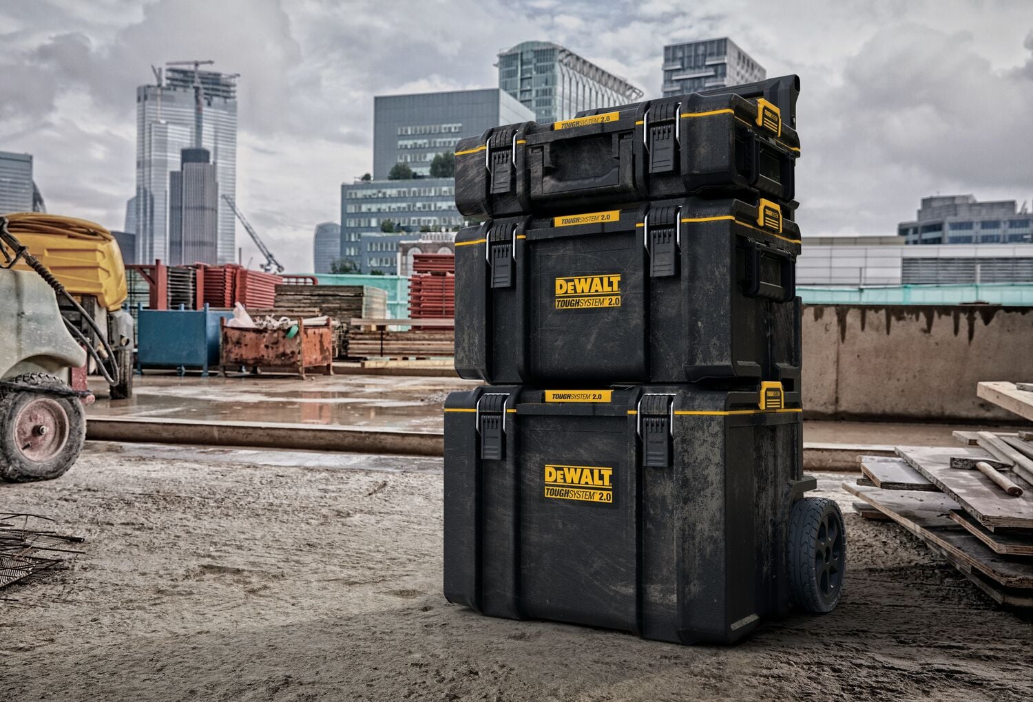 Pila de tres cajas de almacenamiento de herramientas DEWALT ToughSystem 2.0 con ruedas, mostrada en un sitio de construcción con edificios modernos al fondo.